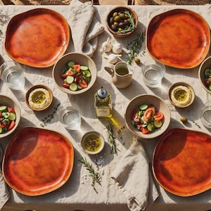 Peut inclure: Un service de table composé de quatre grandes assiettes irrégulières orange brûlé et quatre bols beiges assortis. La table comprend des verres transparents, des petits bols de houmous, une burette à huile en verre et divers aliments frais comme du pain tranché, une salade de tomates et concombres, des olives et des gousses d'ail disposés sur une nappe en lin clair.