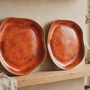 Può includere: Due piatti in ceramica con una forma organica irregolare e una vivace smaltatura arancione maculata. La superficie di ogni piatto presenta una finitura lucida con toni di rosso e marrone più scuri mescolati al colore arancione brillante.