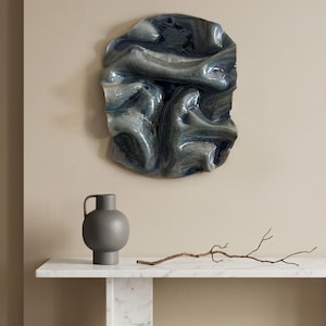 May include: An interior scene with a wall-mounted abstract artwork in blue and grey tones. A grey vase with a handle is on a white marble shelf, with a dried branch. The wall is painted a neutral beige. The vase is approximately 20 cm tall.