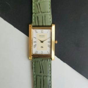 May include: A rectangular gold-toned Seiko Quartz watch with a white face and Roman numeral hour markers. The watch has gold hands and a green leather strap. The strap has a textured, crocodile-like pattern.