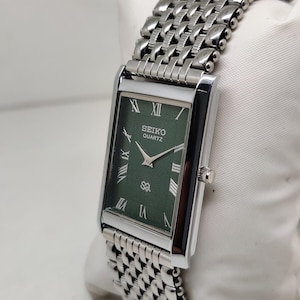 Może przedstawiać: Prostokątny zegarek Seiko Quartz ze srebrną metalową bransoletą i kopertą. Tarcza zegarka jest w kolorze ciemnozielonym ze srebrnymi wskaz&oacute;wkami i rzymskimi cyframi. Zegarek jest prezentowany na białej powierzchni.