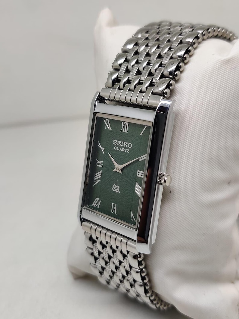 Może przedstawiać: Prostokątny zegarek Seiko Quartz ze srebrną metalową bransoletą i kopertą. Tarcza zegarka jest w kolorze ciemnozielonym ze srebrnymi wskaz&oacute;wkami i rzymskimi cyframi. Zegarek jest prezentowany na białej powierzchni.