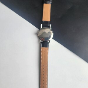 Puede incluir: Un reloj de pulsera plateado con una correa de cuero negro y una parte inferior marr&oacute;n claro. El reloj tiene una esfera redonda y un cierre de hebilla. La correa est&aacute; sujeta a la caja del reloj con metal plateado. El reloj se muestra sobre un fondo blanco y negro.