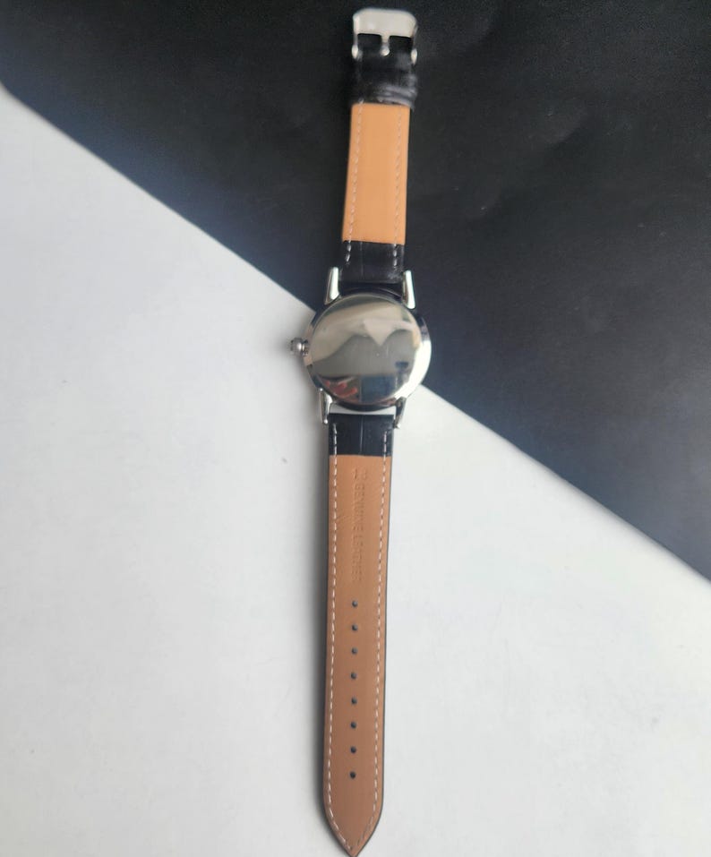 Puede incluir: Un reloj de pulsera plateado con una correa de cuero negro y una parte inferior marr&oacute;n claro. El reloj tiene una esfera redonda y un cierre de hebilla. La correa est&aacute; sujeta a la caja del reloj con metal plateado. El reloj se muestra sobre un fondo blanco y negro.