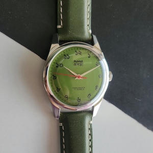 May include: A vintage wristwatch with a green face and a silver case. The watch has a green leather band with white stitching. The watch face displays the time with Arabic numerals and the text "PARA SHOCK 17 JEWELS".