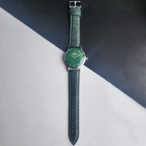 May include: A wristwatch with a green face and a silver-colored case. The watch has a dark green leather band with a silver buckle. The watch is displayed against a black and white background.