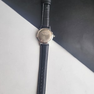 Puede incluir: Un reloj de pulsera Piaget plateado con una correa de cuero negro. La esfera del reloj es redonda con agujas plateadas y una caja plateada. La correa negra tiene costuras blancas y una hebilla plateada.