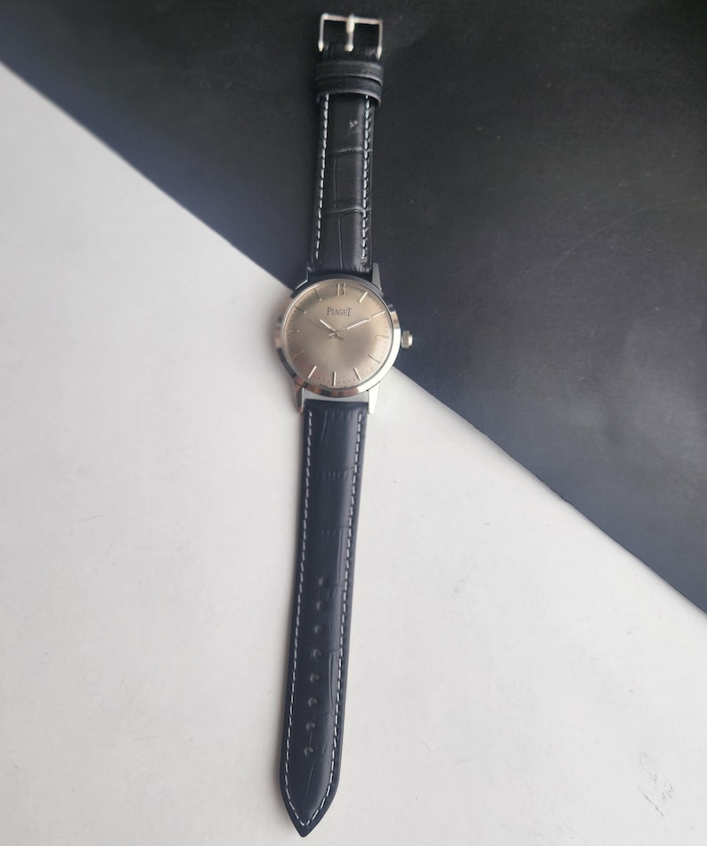 Puede incluir: Un reloj de pulsera Piaget plateado con una correa de cuero negro. La esfera del reloj es redonda con agujas plateadas y una caja plateada. La correa negra tiene costuras blancas y una hebilla plateada.