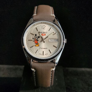 Op de afbeelding: Zilverkleurig Seiko 5 automatisch horloge met een lichtgekleurde wijzerplaat, met een Mickey Mouse-afbeelding. Het horloge heeft een bruine leren band en geeft de dag en datum weer. De wijzerplaat heeft de tekst "Seiko 5 Automatic Japan Made".