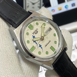May include: A stainless steel Seiko automatic wristwatch with a cream-colored face. The watch face features green and blue accents, a day-date display, and a black leather strap. The watch hands and hour markers glow in the dark.