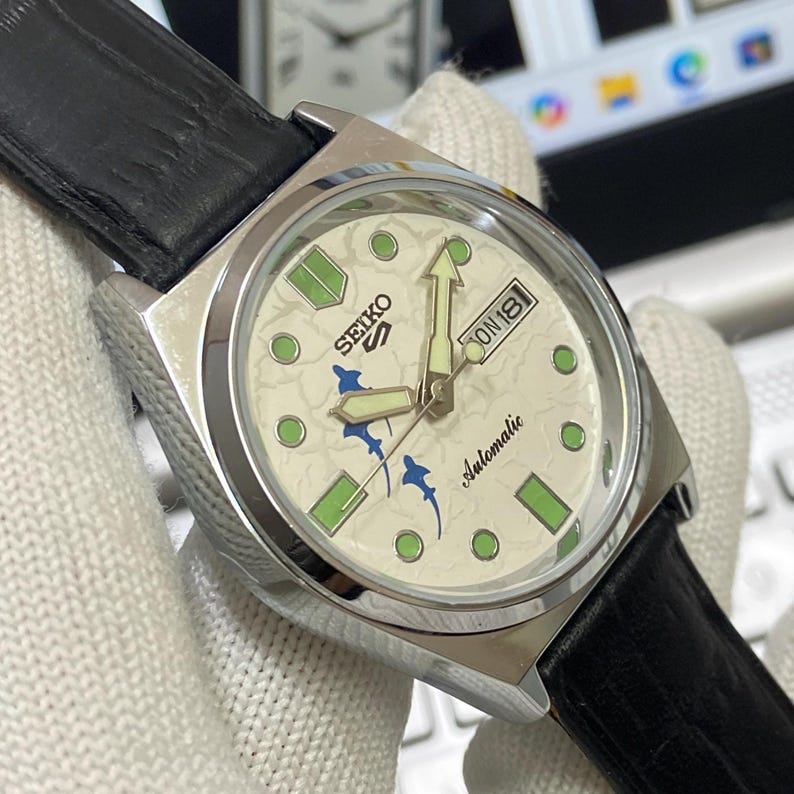 May include: A stainless steel Seiko automatic wristwatch with a cream-colored face. The watch face features green and blue accents, a day-date display, and a black leather strap. The watch hands and hour markers glow in the dark.