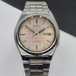 May include: A silver-toned Seiko 5 automatic wristwatch with a pinkish-beige dial. The watch face features a grid pattern, day-date display, and silver hands and markers. The watch band is made of silver-toned metal links.