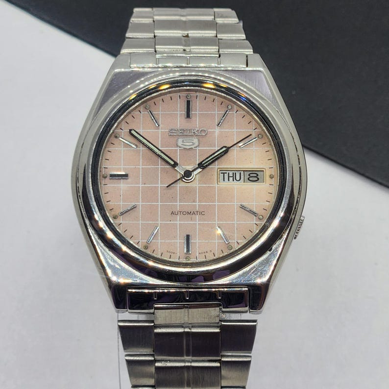 May include: A silver-toned Seiko 5 automatic wristwatch with a pinkish-beige dial. The watch face features a grid pattern, day-date display, and silver hands and markers. The watch band is made of silver-toned metal links.