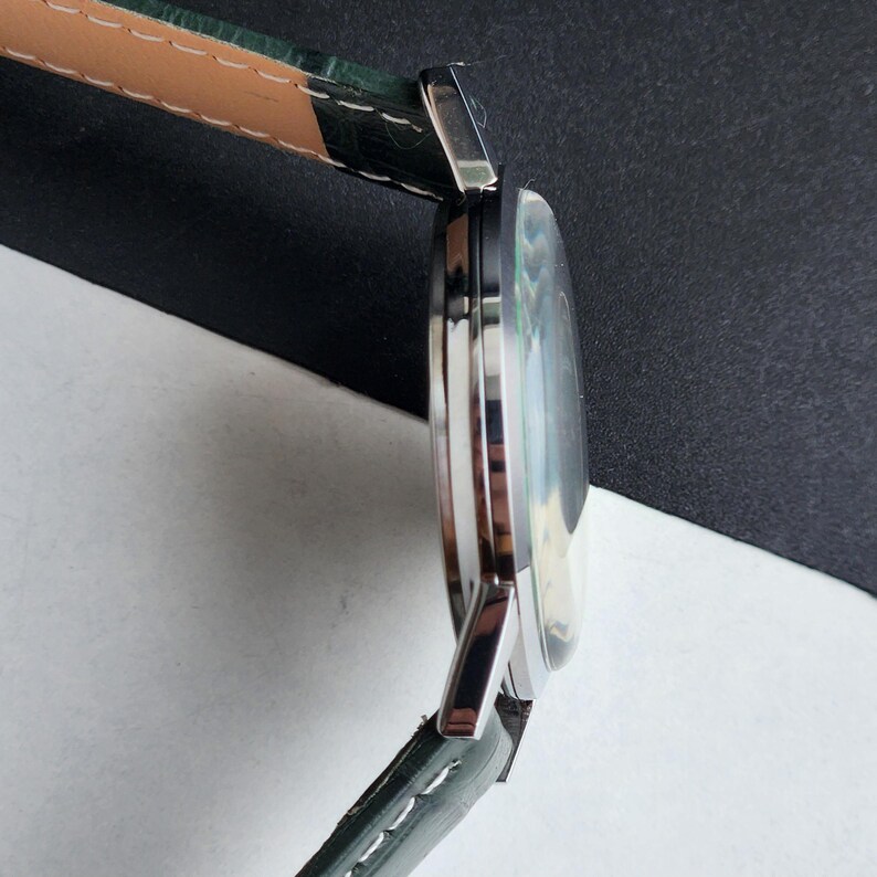 May include: A silver wristwatch with a round face and a green leather band. The watch face has a light-colored, iridescent appearance. The band is dark green with a lighter tan underside and white stitching.