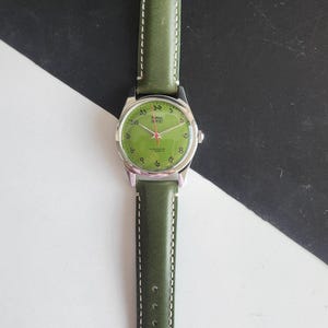 May include: A wristwatch with a green leather band and a silver-colored case. The watch face is green with black numerals and hands. The band is stitched with white thread.