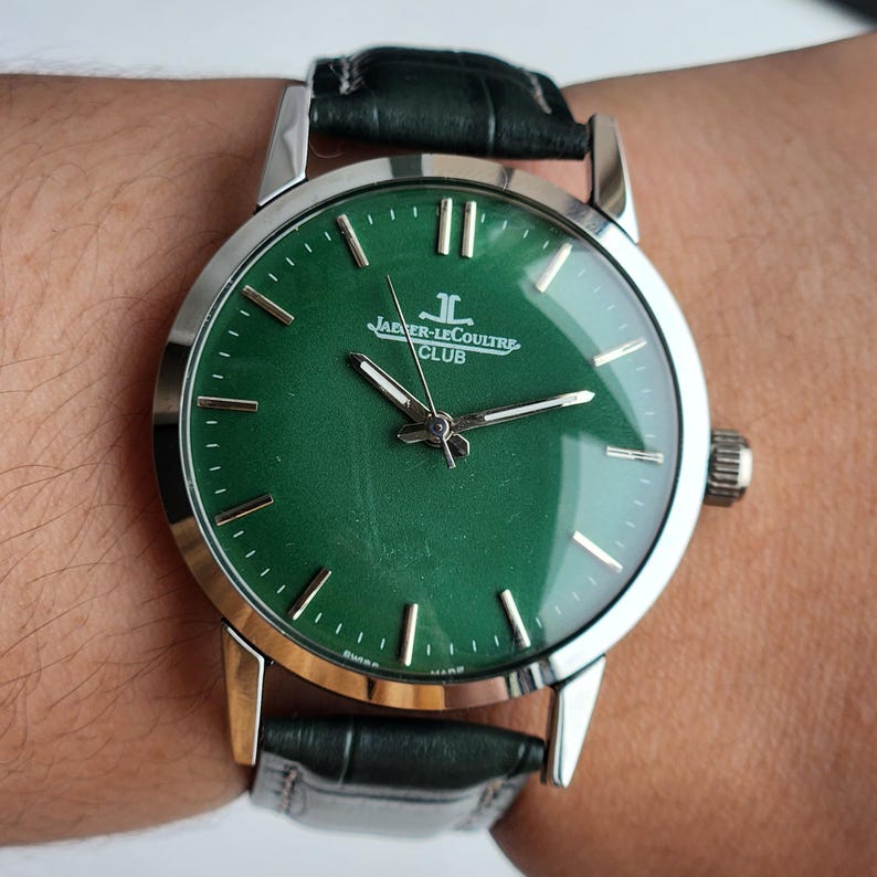 May include: A wristwatch with a green face and silver-colored case. The watch has a black leather band and the words "JAEGER-LECOULTRE CLUB" are printed on the face. The watch is on a wrist.