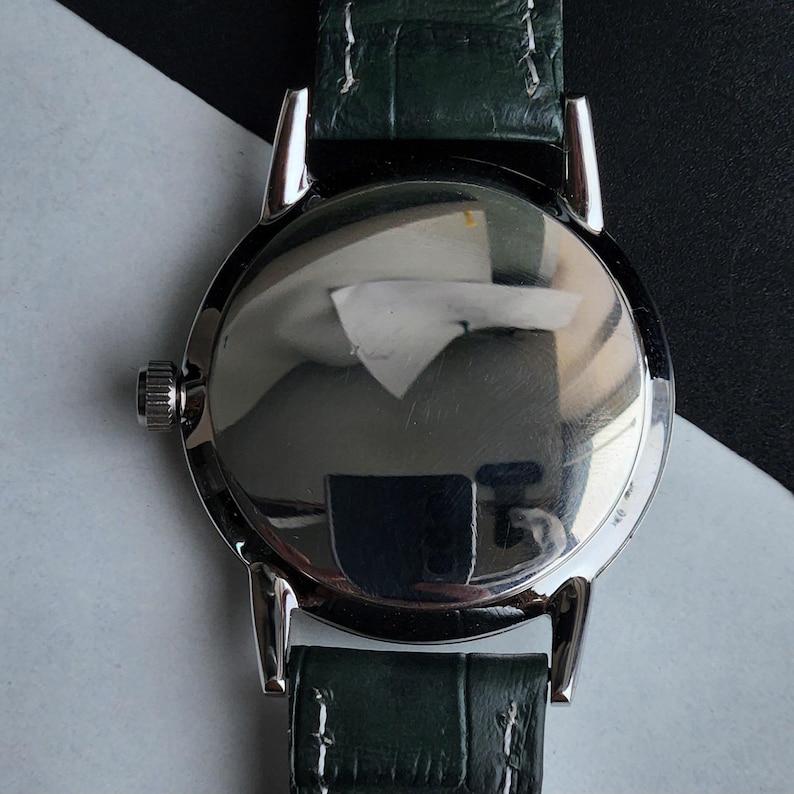 May include: Close-up of the back of a silver wristwatch with a dark green leather band. The watch face is reflective, showing a distorted view of the surroundings. The band is textured and appears to be made of genuine leather.