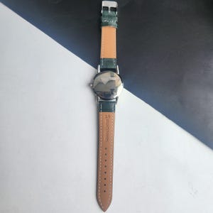 May include: A silver-toned wristwatch with a green leather band and a light brown underside. The watch face is reflective, and the band is secured with a silver buckle. The words "GENUINE LEATHER" are printed on the band.