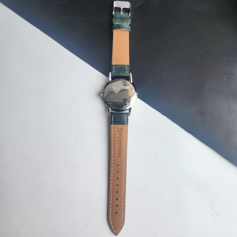 May include: A silver-toned wristwatch with a green leather band and a light brown underside. The watch face is reflective, and the band is secured with a silver buckle. The words "GENUINE LEATHER" are printed on the band.