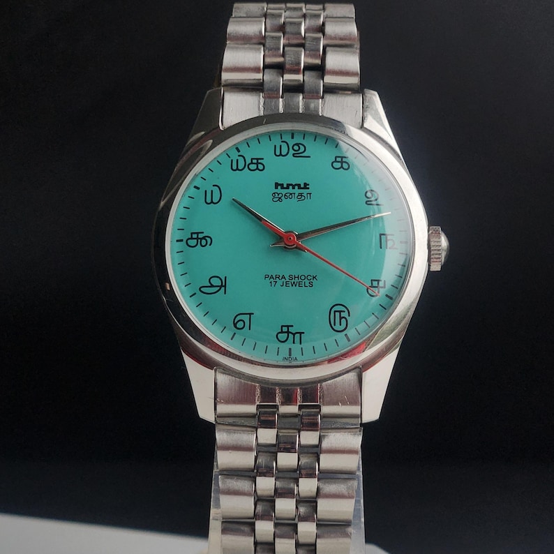 May include: A silver-toned wristwatch with a teal face and a matching silver band. The watch face features black text and numerals, with red second hand. The watch is labeled "PARA SHOCK 17 JEWELS".