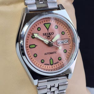 K&ouml;nnte beinhalten: Eine Seiko 5 Automatik-Uhr aus Edelstahl mit einem lachsfarbenen Zifferblatt. Die Uhr verf&uuml;gt &uuml;ber eine Tages- und Datumsanzeige, Leuchtzeiger und gr&uuml;ne Stundenmarkierungen. Das Armband ist silberfarben und mehrgliedrig.