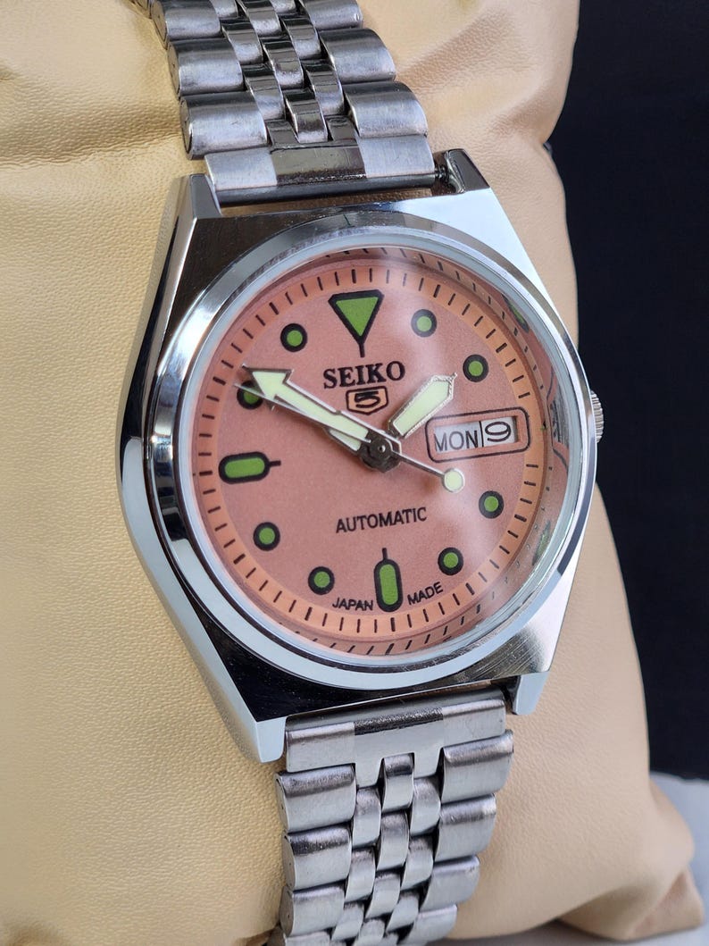 K&ouml;nnte beinhalten: Eine Seiko 5 Automatik-Uhr aus Edelstahl mit einem lachsfarbenen Zifferblatt. Die Uhr verf&uuml;gt &uuml;ber eine Tages- und Datumsanzeige, Leuchtzeiger und gr&uuml;ne Stundenmarkierungen. Das Armband ist silberfarben und mehrgliedrig.