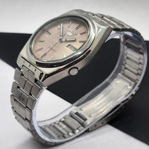 May include: A silver-toned Seiko 5 automatic watch with a light pink dial and a matching silver band. The watch face displays the day and date. The watch is on a clear plastic display stand.