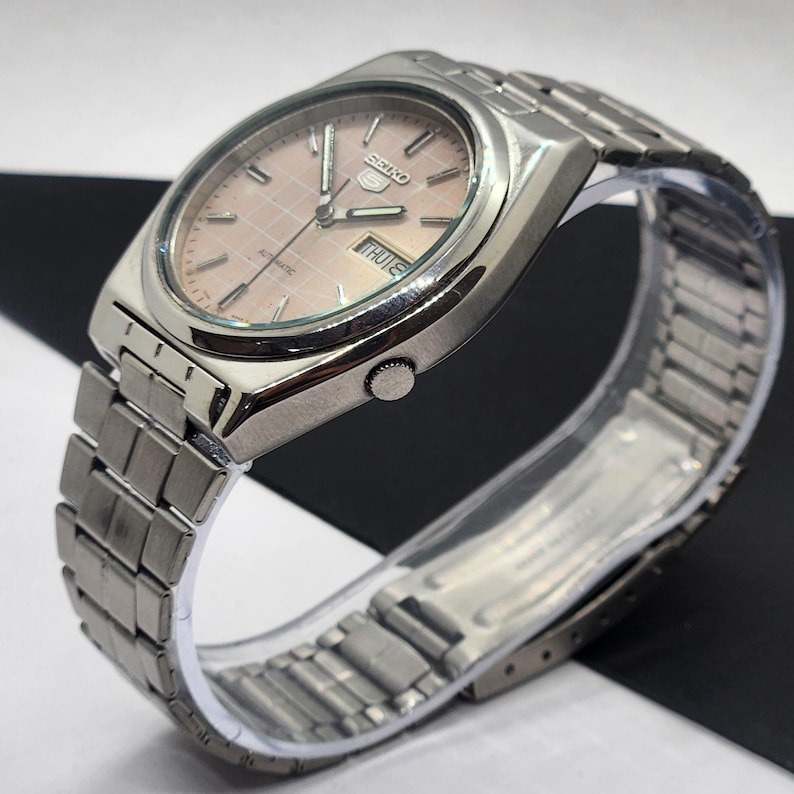 May include: A silver-toned Seiko 5 automatic watch with a light pink dial and a matching silver band. The watch face displays the day and date. The watch is on a clear plastic display stand.
