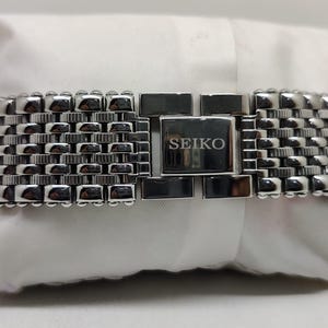 Może przedstawiać: Srebrny pasek do zegarka Seiko z tkanym, teksturowanym wzorem. Pasek posiada prostokątne zapięcie z logo Seiko. Pasek do zegarka spoczywa na białej powierzchni.