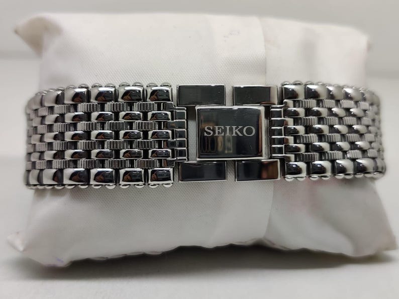 Może przedstawiać: Srebrny pasek do zegarka Seiko z tkanym, teksturowanym wzorem. Pasek posiada prostokątne zapięcie z logo Seiko. Pasek do zegarka spoczywa na białej powierzchni.
