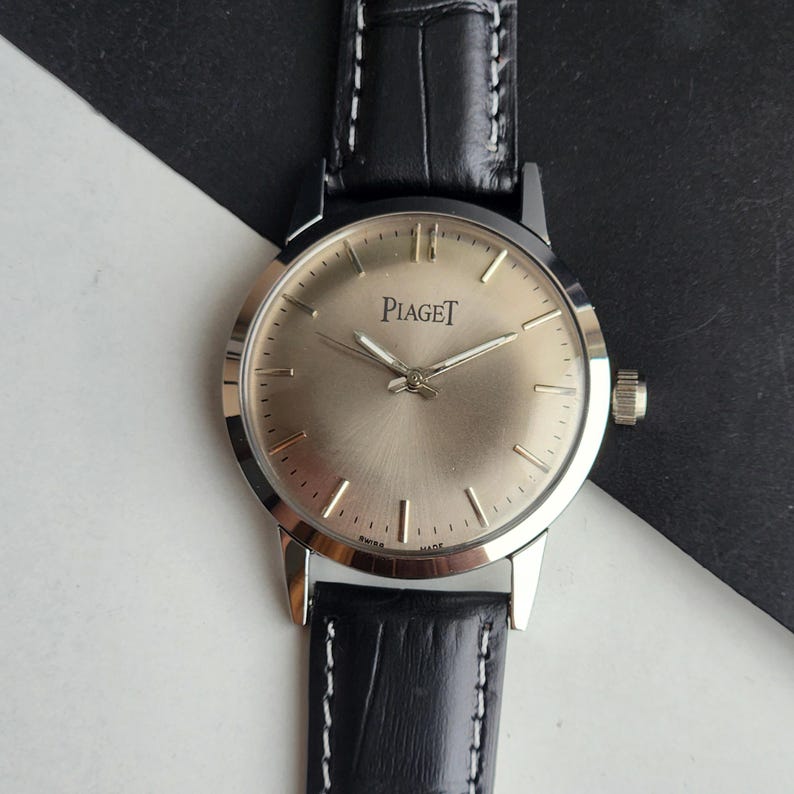 Puede incluir: Un reloj Piaget plateado con esfera dorada clara y correa de cuero negro. El reloj presenta agujas e indicadores de hora plateados, y el nombre de la marca se muestra de forma destacada. La correa tiene costuras blancas.