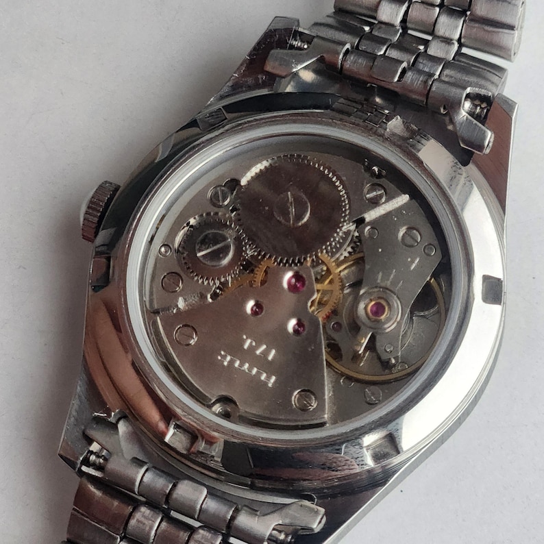 Puede incluir: Primer plano de un reloj de pulsera vintage con una correa de metal plateado. La parte trasera del reloj est&aacute; abierta, revelando el intrincado movimiento mec&aacute;nico con engranajes, joyas y el texto "hmt: 17J."