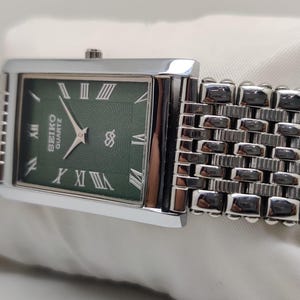 Może przedstawiać: Prostokątny zegarek Seiko Quartz z zieloną tarczą i srebrnym paskiem. Tarcza zegarka posiada rzymskie cyfry i srebrne wskaz&oacute;wki. Pasek zegarka jest w stylu tkanym. Zegarek ma klasyczny design.