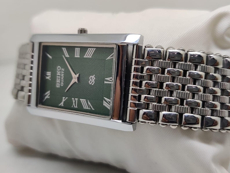 Może przedstawiać: Prostokątny zegarek Seiko Quartz z zieloną tarczą i srebrnym paskiem. Tarcza zegarka posiada rzymskie cyfry i srebrne wskaz&oacute;wki. Pasek zegarka jest w stylu tkanym. Zegarek ma klasyczny design.