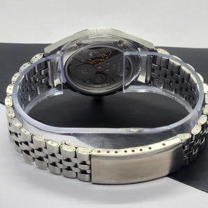 May include: A silver-toned wristwatch with a transparent case back, revealing the intricate mechanical movement. The watch features a stainless steel band with a clasp. The watch is displayed on a clear plastic holder.