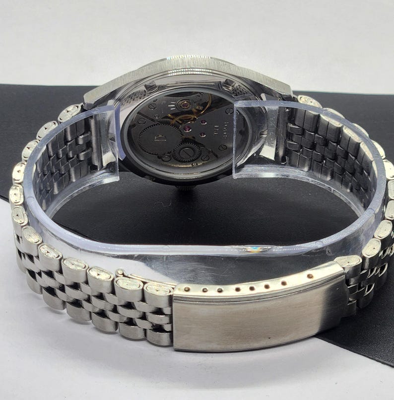 May include: A silver-toned wristwatch with a transparent case back, revealing the intricate mechanical movement. The watch features a stainless steel band with a clasp. The watch is displayed on a clear plastic holder.