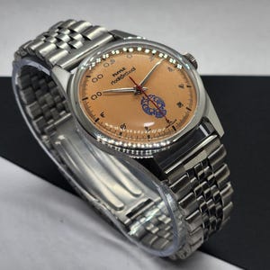 May include: A vintage wristwatch with a stainless steel band and case. The watch face is a light orange color with black numerals and hands. The watch face also features the text "hmt roorkee school" and a blue emblem. The watch is displayed on a clear plastic holder.