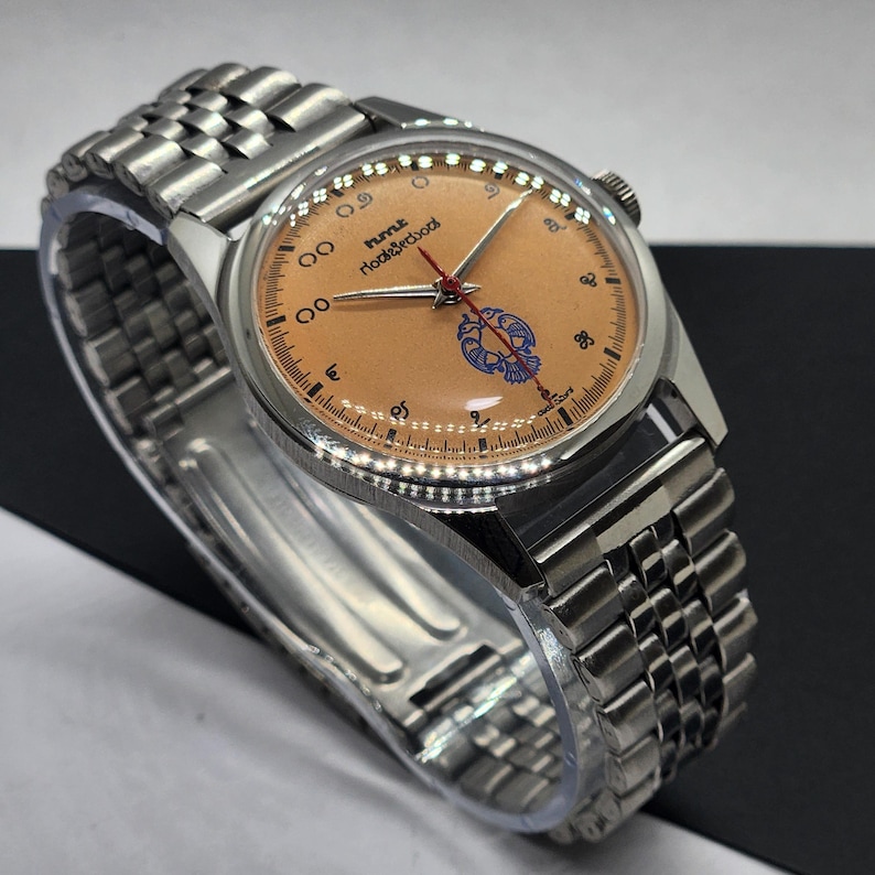May include: A vintage wristwatch with a stainless steel band and case. The watch face is a light orange color with black numerals and hands. The watch face also features the text "hmt roorkee school" and a blue emblem. The watch is displayed on a clear plastic holder.