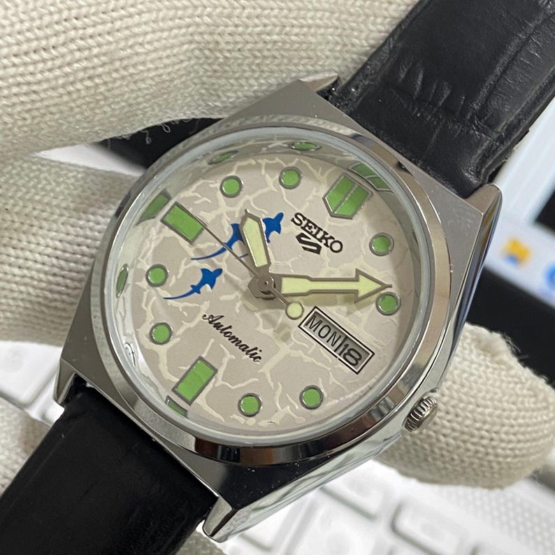 May include: A silver-toned Seiko 5 automatic watch with a patterned light gray face. The watch features green and blue accents, a black leather band, and the day and date displayed at the 3 o'clock position.
