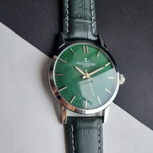 May include: A close-up of a Jaeger-LeCoultre Club watch with a green face and silver casing. The watch has gold hands and markers, and a dark green leather band. The watch is set against a black and white background.
