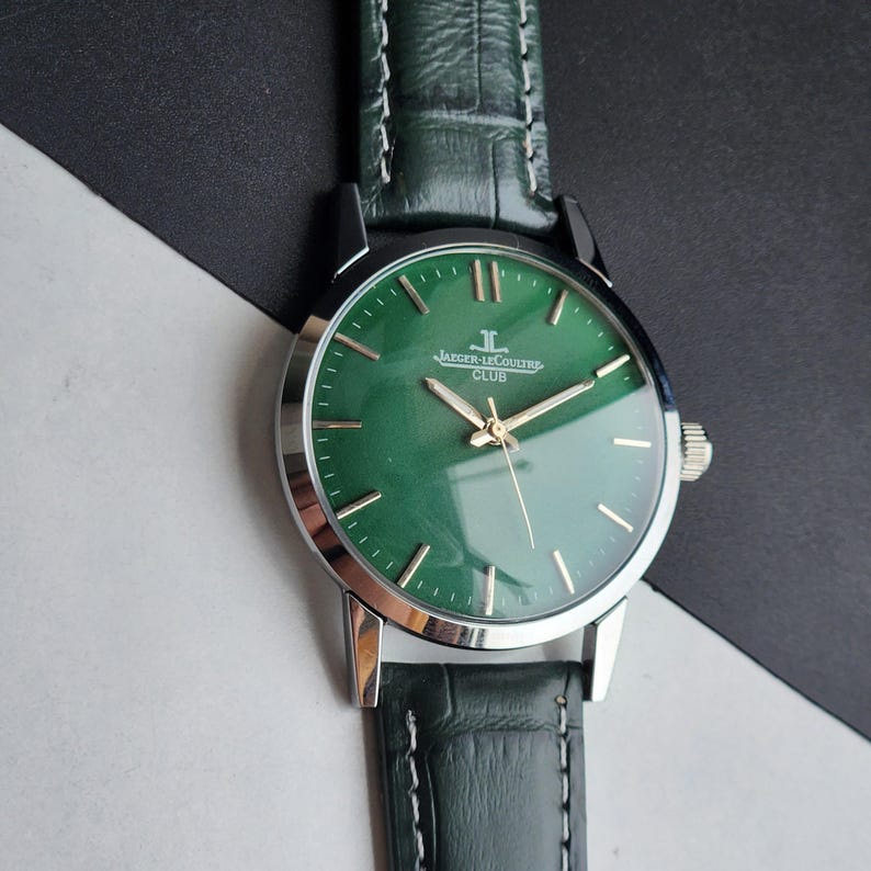 May include: A close-up of a Jaeger-LeCoultre Club watch with a green face and silver casing. The watch has gold hands and markers, and a dark green leather band. The watch is set against a black and white background.
