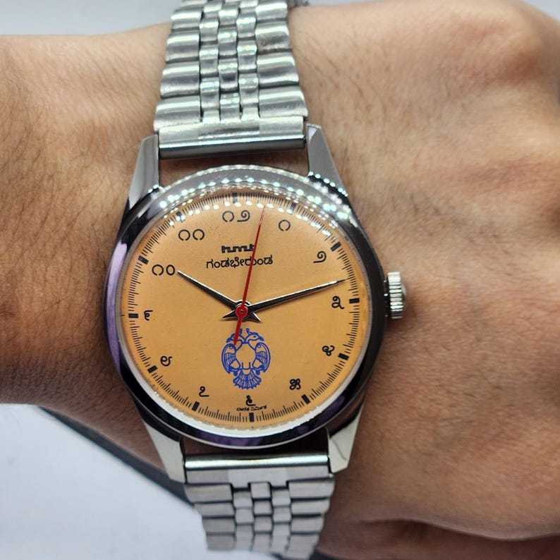 May include: A silver wristwatch with a light orange face and a silver band. The watch face has black numerals, a red second hand, and text in a foreign language. The watch is on a person's wrist.