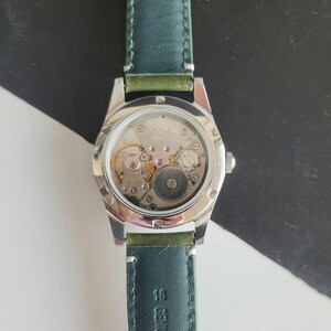 May include: A silver-toned wristwatch with a clear back, showcasing the intricate internal mechanisms. The watch has a dark green leather band. The watch face displays gears, springs, and other components. The watch is a classic timepiece.