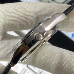 May include: Close-up of a silver wristwatch with a black leather strap. The watch has a polished metal case with a round button and a detailed dial. The watch is set against a white, textured background.