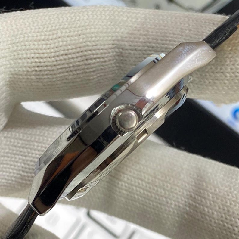 May include: Close-up of a silver wristwatch with a black leather strap. The watch has a polished metal case with a round button and a detailed dial. The watch is set against a white, textured background.