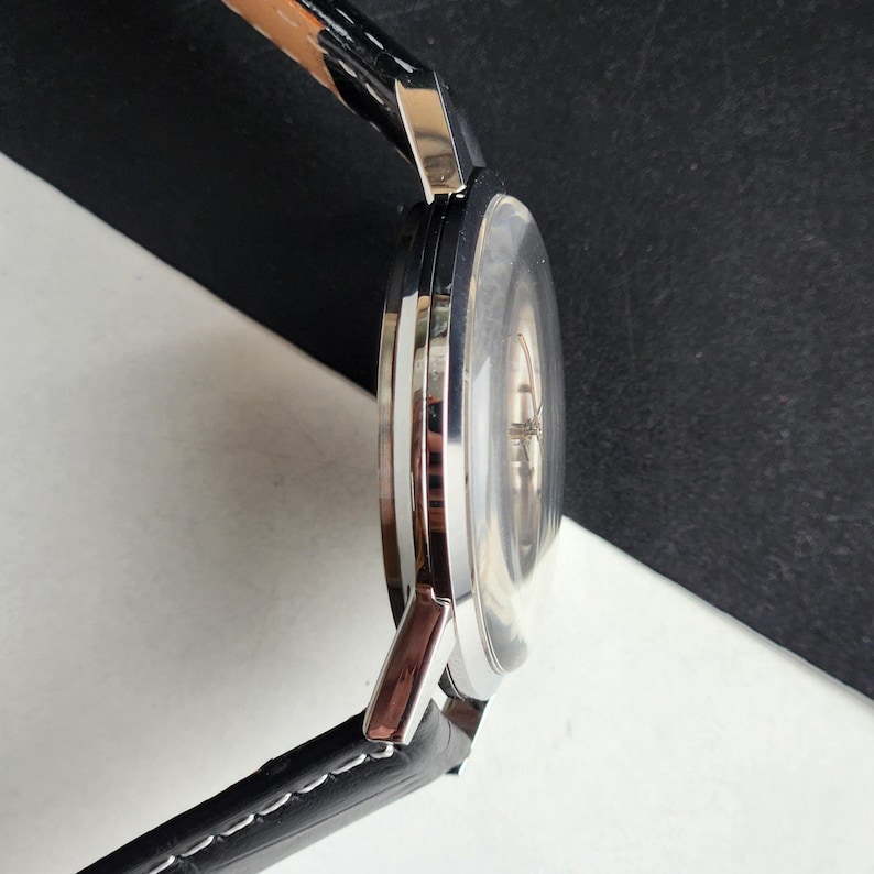 Puede incluir: Vista lateral de un reloj de pulsera plateado con una correa de cuero negro. La esfera del reloj es transparente y la correa est&aacute; cosida con hilo blanco. El reloj tiene un dise&ntilde;o cl&aacute;sico y minimalista.