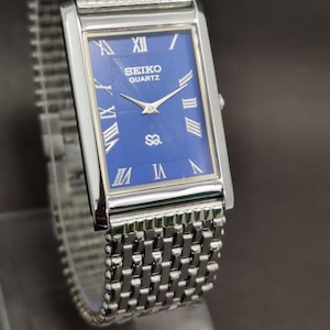 Puede incluir: Un reloj Seiko Quartz rectangular con esfera azul y correa plateada. La esfera del reloj presenta números romanos y agujas plateadas. La correa es de estilo malla. El reloj se exhibe sobre un soporte transparente.