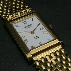Op de afbeelding: Een goudkleurig Seiko Quartz horloge met een rechthoekige wijzerplaat en een geweven metalen band. De wijzerplaat is wit met zwarte Romeinse cijfers en gouden wijzers. Klassiek ontwerp.