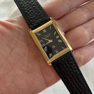 May include: A rectangular gold-toned Seiko Quartz watch with a black face and Roman numeral hour markers. The watch has a black leather strap. The watch face displays the Seiko Quartz logo.