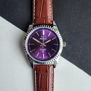 May include: A vintage wristwatch with a purple face and silver case. The watch has gold hands and markers. The brown leather strap is textured. The watch face reads "HMT Kohinoor" and "Para Shock 17 Jewels".